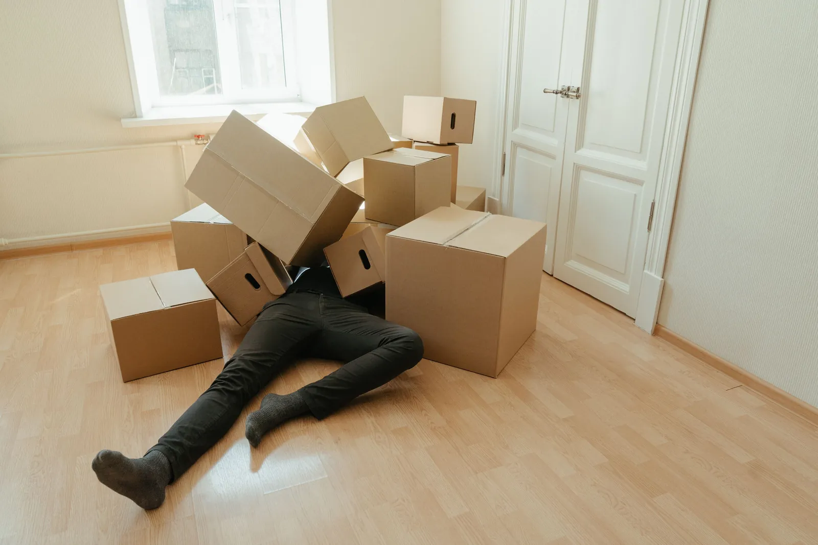 Mann liegt erschöpft unter einem Stapel Umzugskartons auf dem Boden einer leeren Wohnung